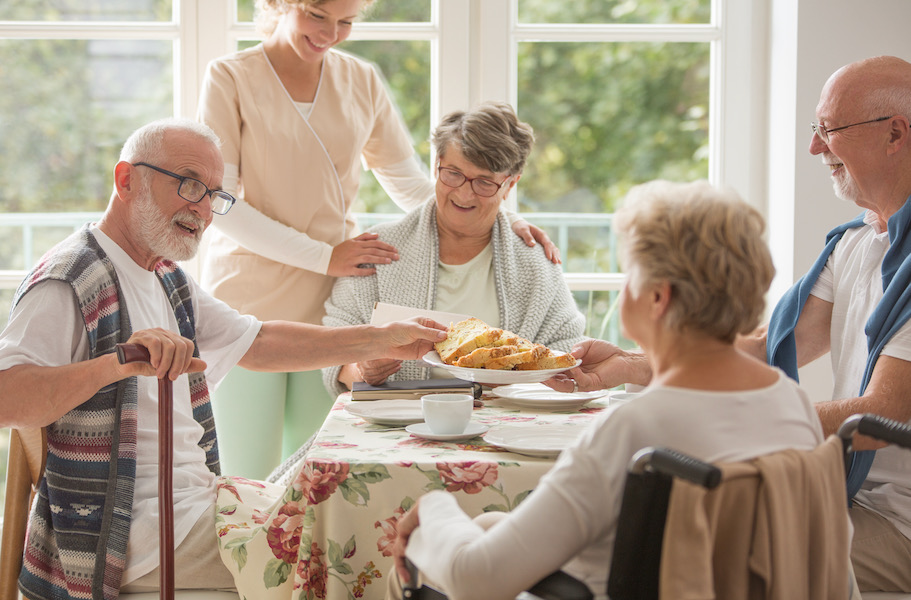 Group-of-senior-friends-with-helpful-carer-sitting-together-at-the-table-at-nursing-home-dining-room-and-eating-cake
